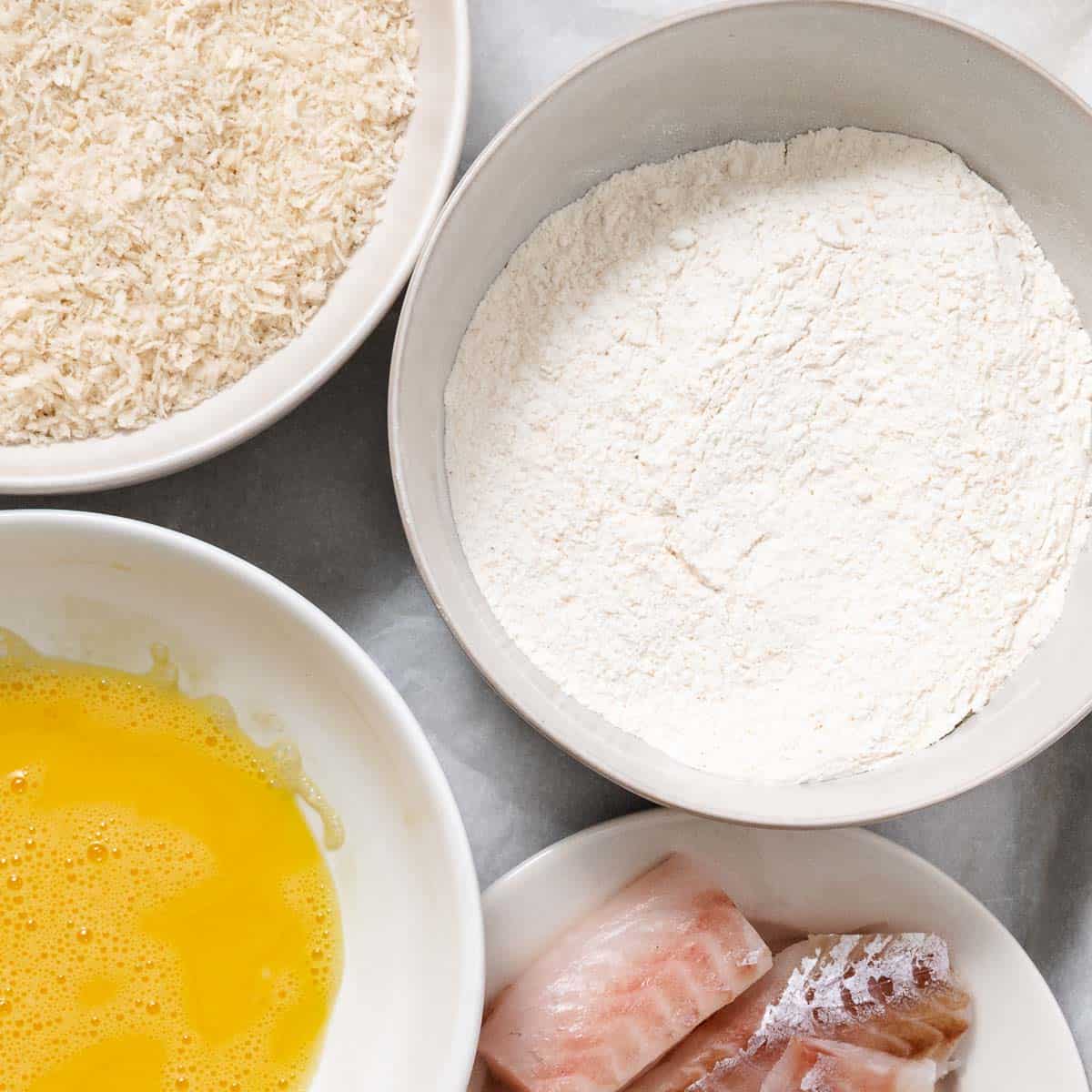Panko, seasoned flour, beaten eggs, and raw pollock set up for breading.