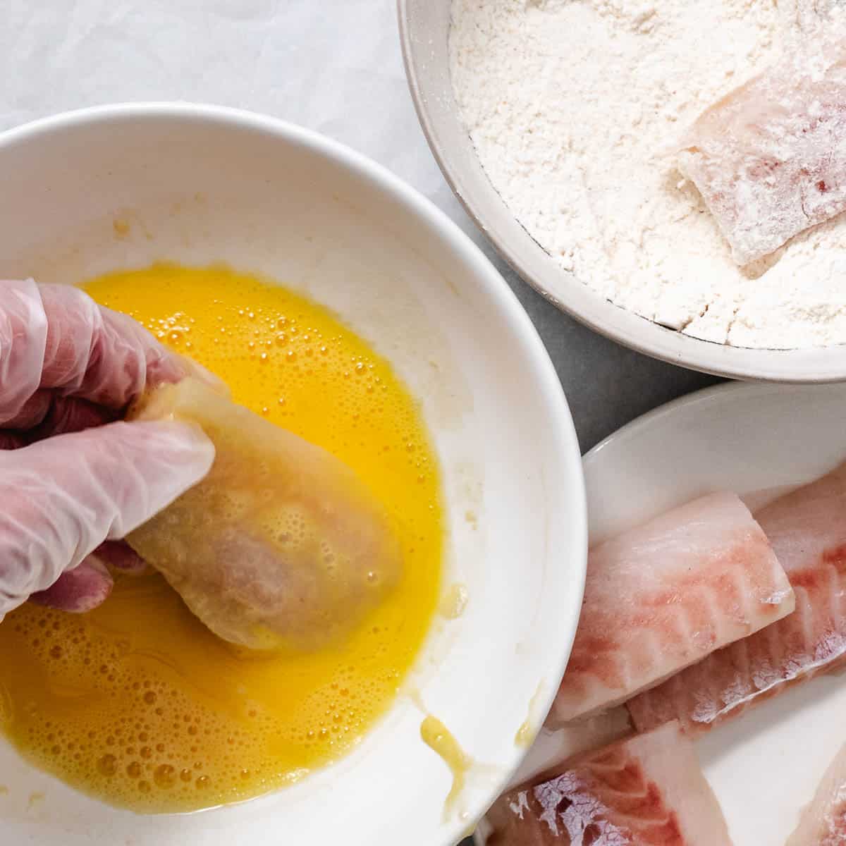 Gloved hand dipping a flour-coated pollock fillet in beaten egg, with a bowl of seasoned flour and more raw pollock nearby.