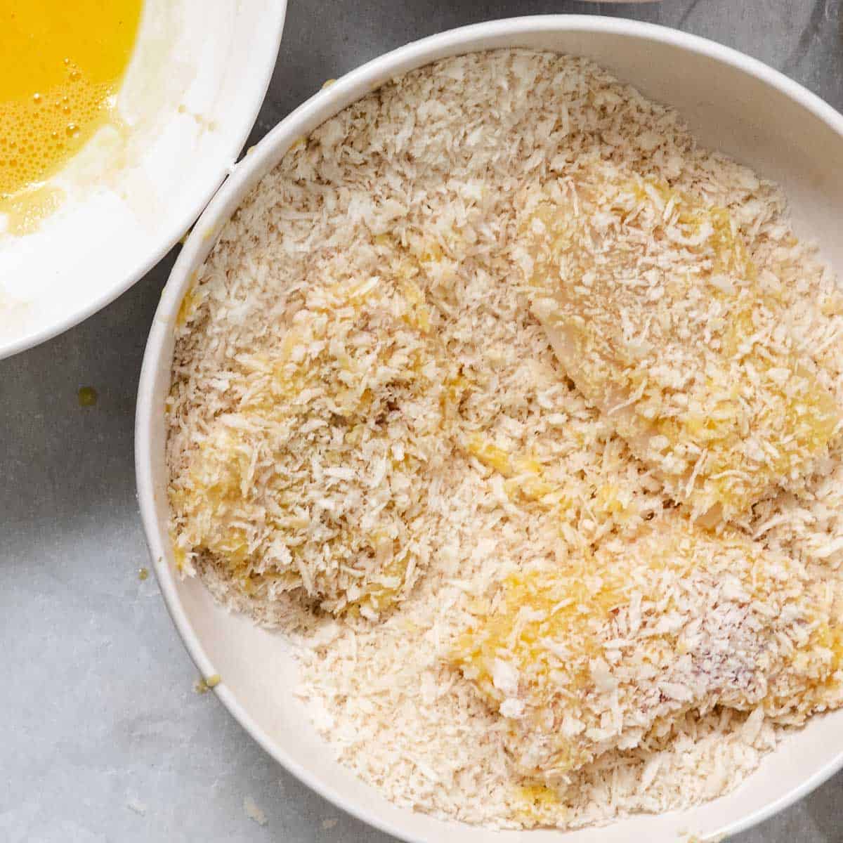 Pollock fillets coated in panko breadcrumbs in a shallow white bowl, with beaten egg alongside.