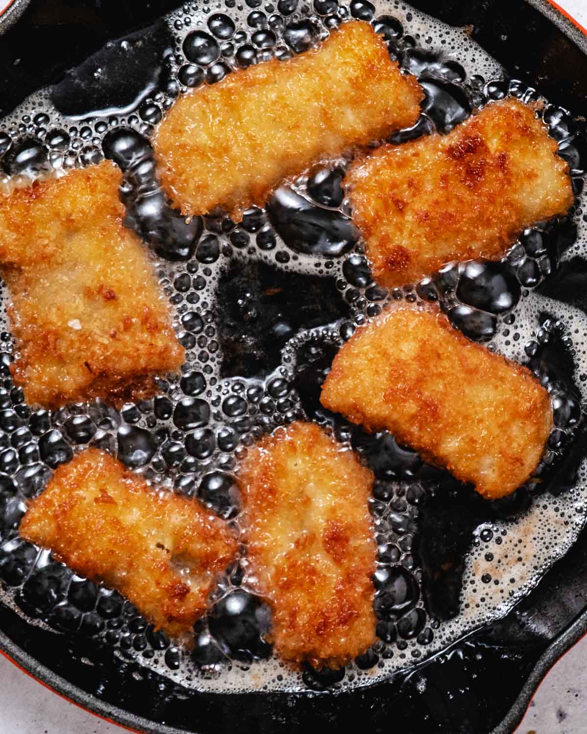 Panko-crusted pollock pieces frying golden brown in bubbling oil in a cast iron skillet.