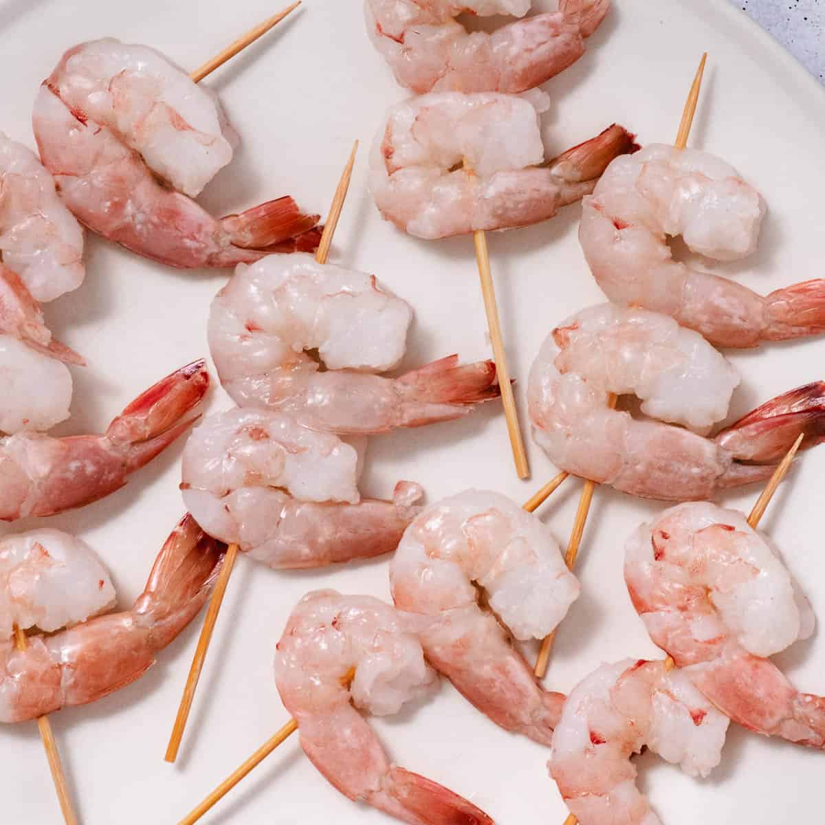Two raw jumbo shrimp threaded onto each short wooden skewer on a white plate.