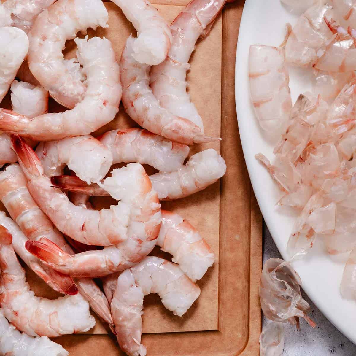 Peeled raw shrimp on a cutting board beside discarded shells.