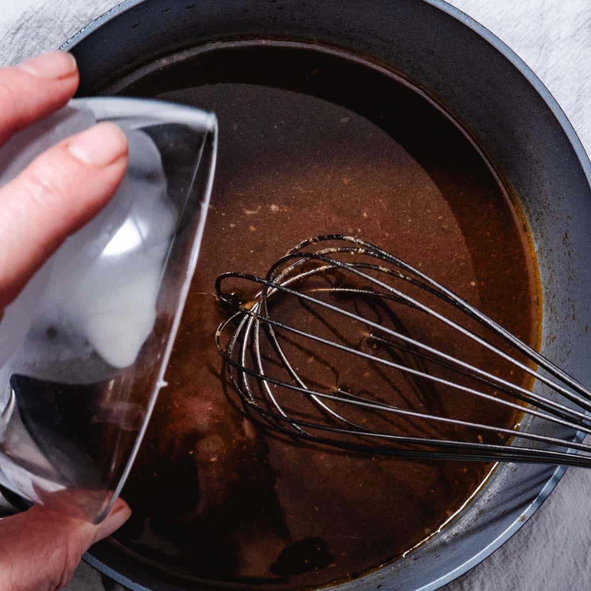 Cornstarch slurry being added to brown cooking liquid in a saucepan with a whisk.