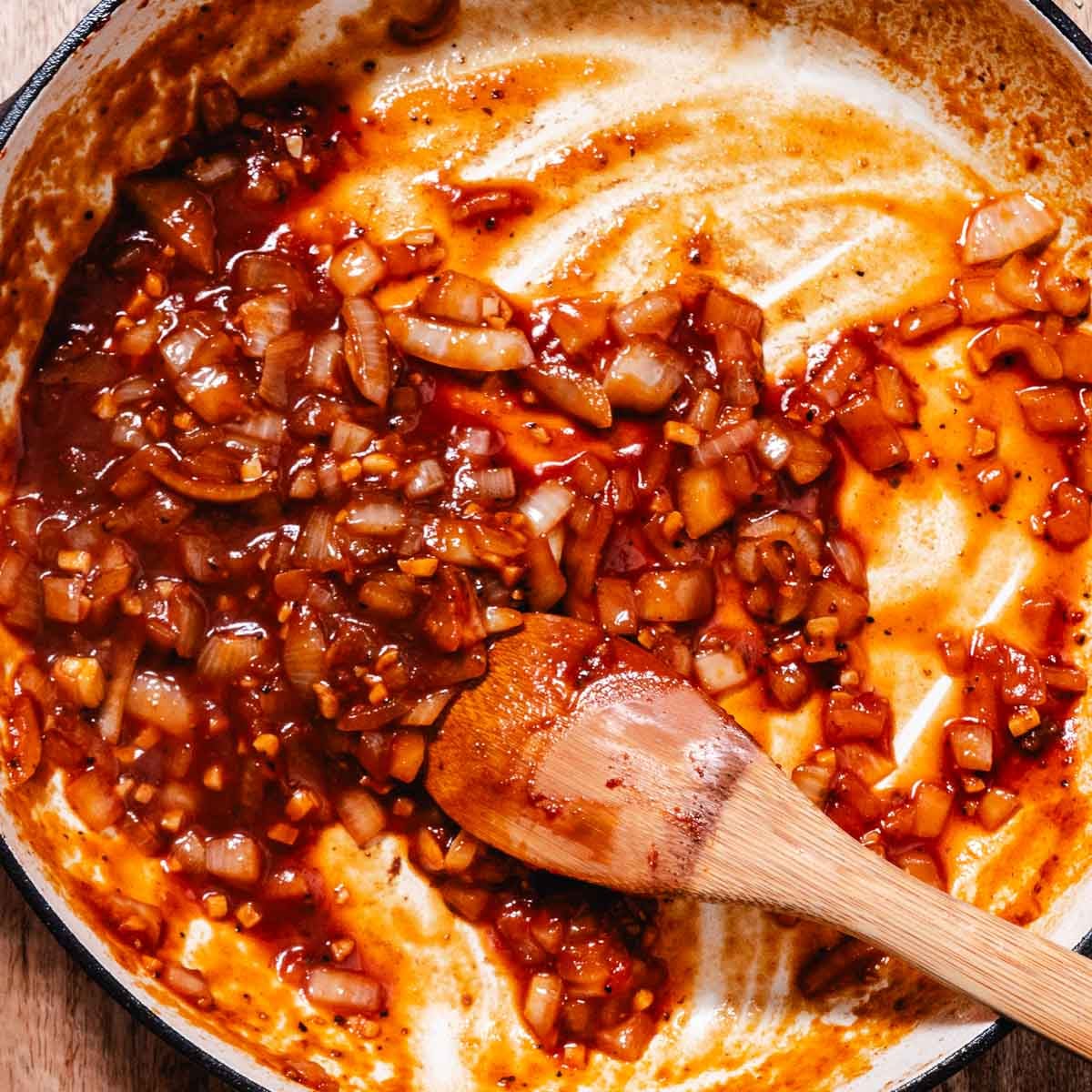 Onions and garlic cooking with tomato paste in a skillet with a wooden spoon.