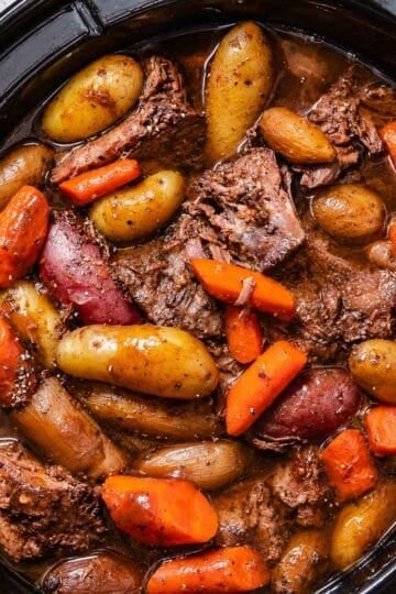 Slow cooker chuck roast with carrots, fingerling potatoes, onions, and brown cooking liquid in a black ceramic crock.