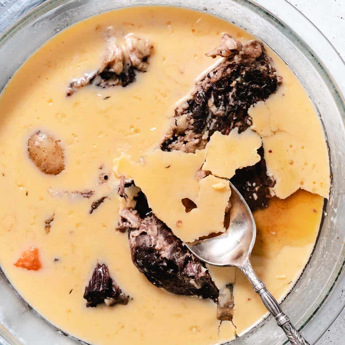 Spoon lifting hardened fat from chilled pot roast and cooking liquid in a glass bowl.