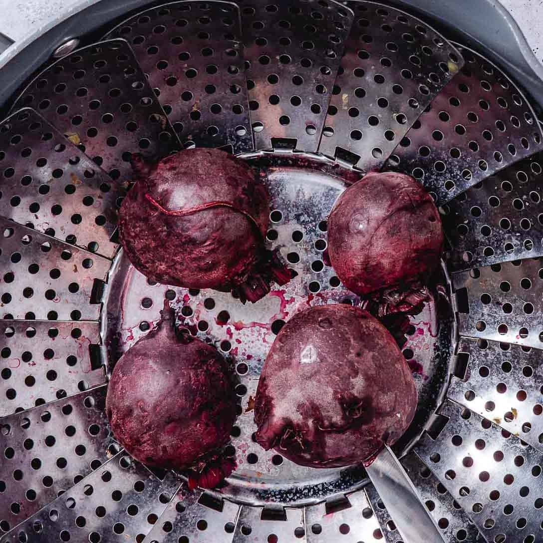 Whole red beets steaming in a covered pot with a steamer basket.